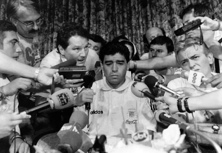 A black-and-white photo of Diego Maradona in an Argentina shirt, sitting amid a crowd of reporters holding microphones and recorders close to his face during a press conference.