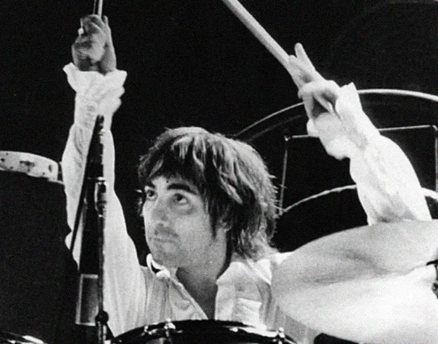 A drummer with shaggy hair and a white shirt energetically plays a drum set, raising both drumsticks above his head on stage under dramatic lighting.