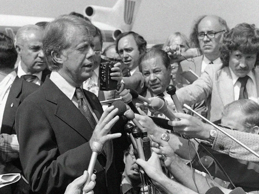 A man in a suit speaks to a large group of reporters holding microphones and recording devices, as photographers and journalists surround him outdoors near an airplane. The image is black and white.
