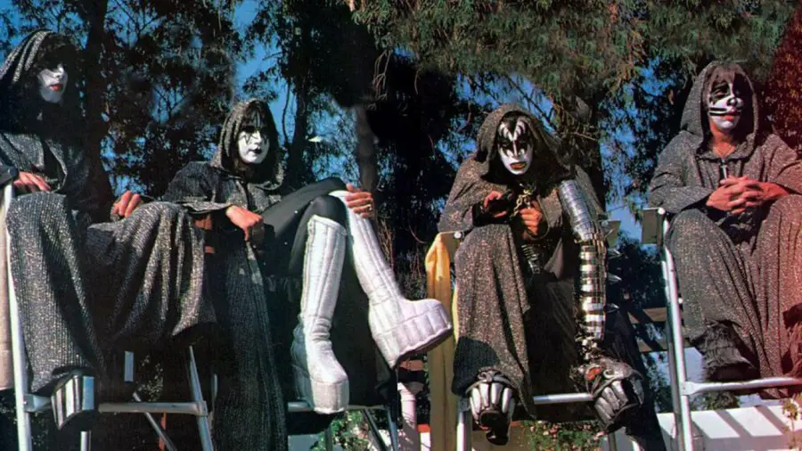 Four people dressed in black hooded robes and wearing dramatic black-and-white face paint sit outdoors on chairs. One has tall platform boots, and another has metallic arm accessories. Trees are visible in the background.