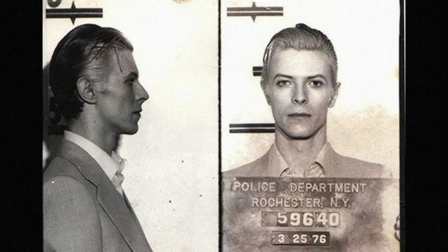 A black-and-white mugshot photo shows a man in a suit with slicked-back hair, facing left in one view and looking forward in another. He holds a placard labeled "Police Department Rochester, N.Y. 3-25-76.