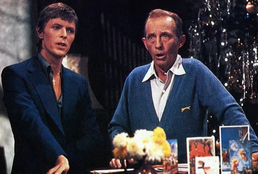 Two men, one younger in a dark suit and one older in a blue sweater, stand side by side singing in front of a Christmas tree and holiday cards.