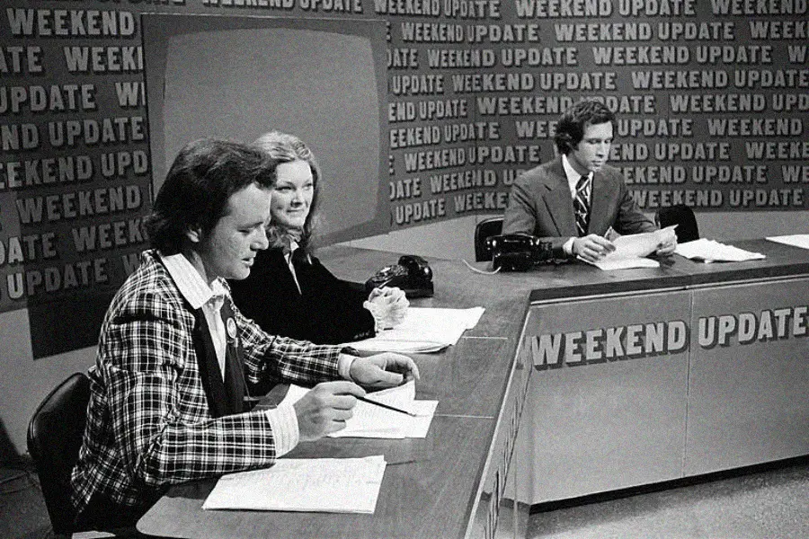 Three people sit at a news desk labeled "WEEKEND UPDATE," reading papers. The background repeats the same text. The image is black and white, suggesting it is from an earlier era.
