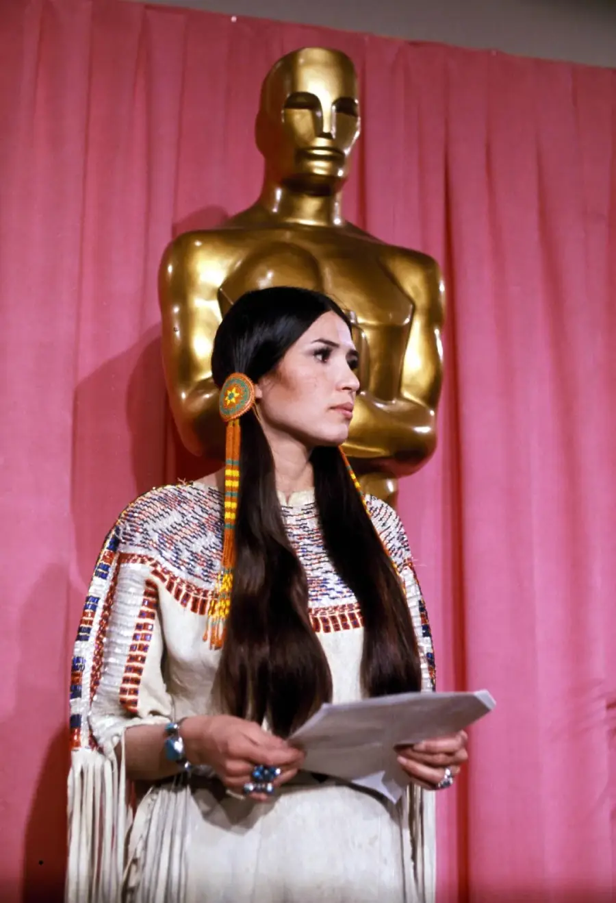 A woman in traditional Native American attire stands holding papers in front of a large gold Oscar statue and a pink curtain background.