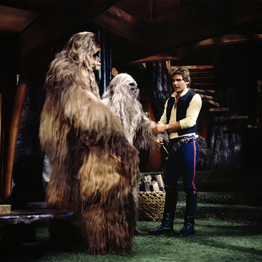 Two tall, furry Wookiee characters stand indoors with a man in a yellow shirt and dark pants. The man is shaking hands with one Wookiee. The setting appears to be a rustic, wooden room with a basket of logs nearby.
