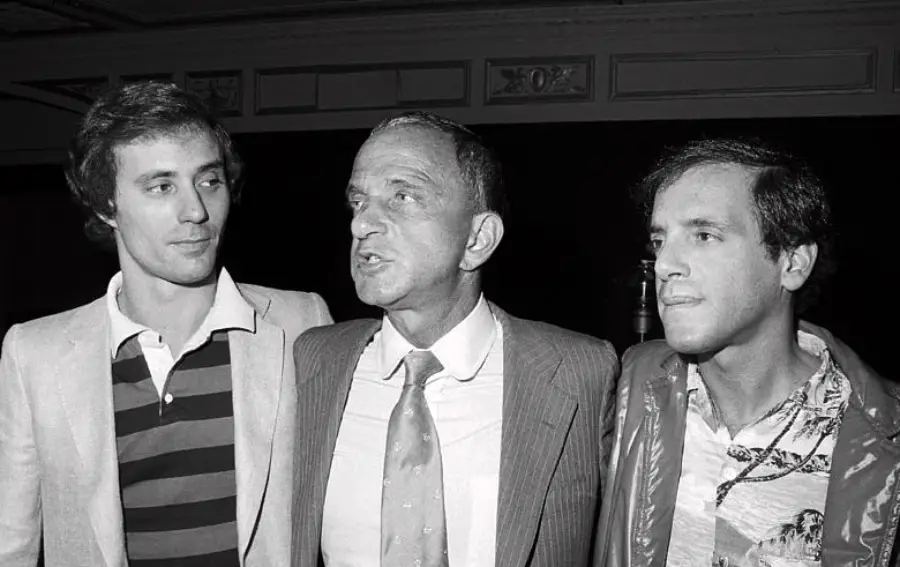 Three men stand together in a black-and-white photo. The man in the middle wears a suit and tie, while the two men on either side wear casual jackets and shirts. All three are looking in different directions.