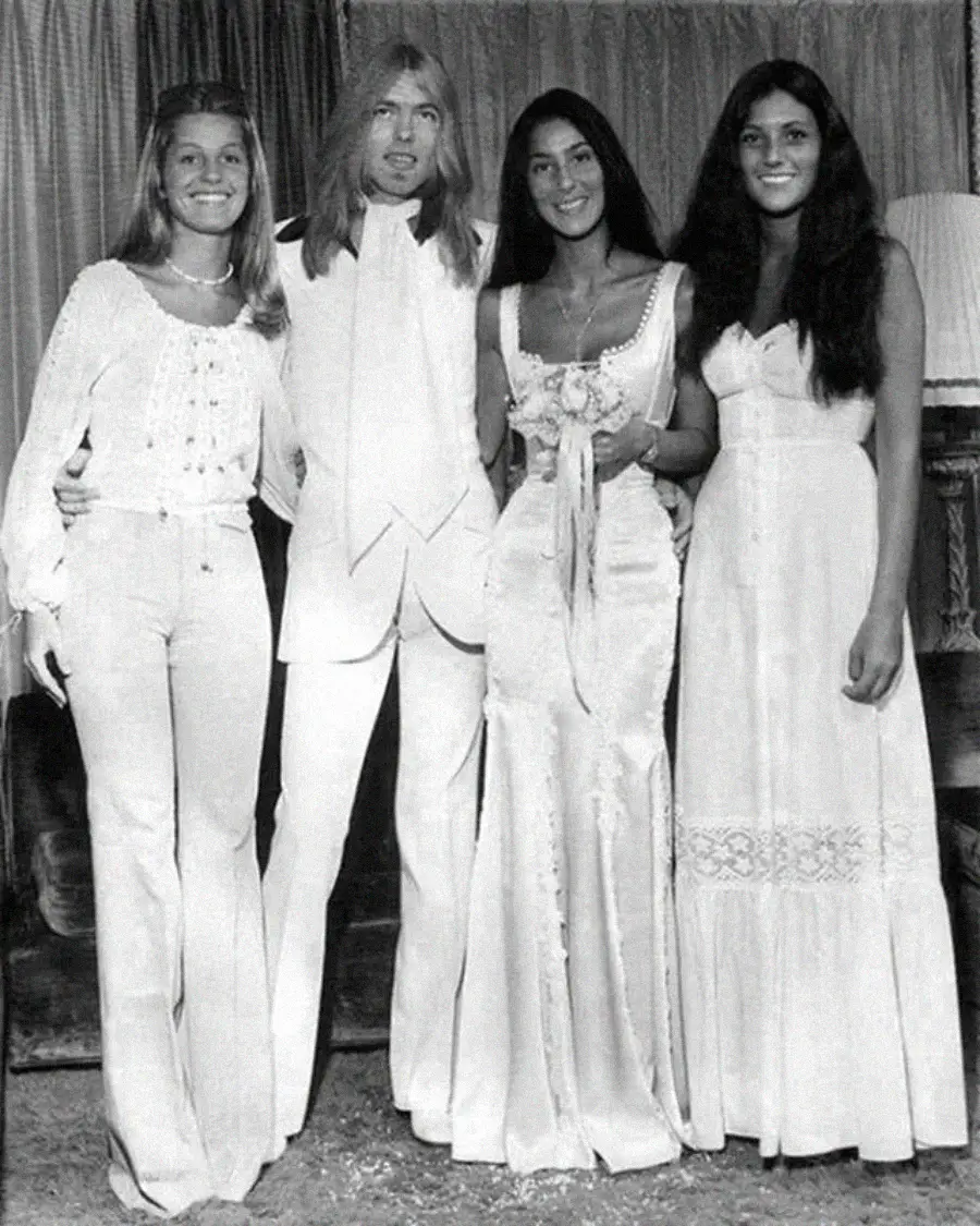 Four people stand together indoors, dressed in formal 1970s-style white clothing; two women wear long dresses, one woman wears pants and a blouse, and a man wears a suit. They are smiling in front of curtains and a lamp.