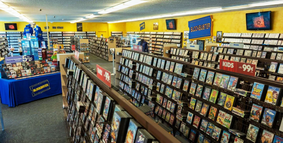 A brightly lit Blockbuster video rental store with rows of DVD and VHS cases, a Kids section sign, and display tables. The store has yellow walls and blue Blockbuster branding throughout.