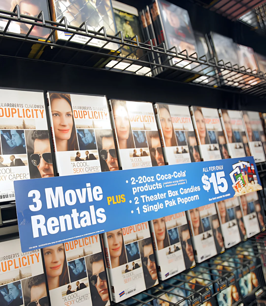 DVD cases of the movie "Duplicity" are displayed on a store shelf. A sign in front advertises a deal: 3 movie rentals plus snacks, drinks, and popcorn for $15.