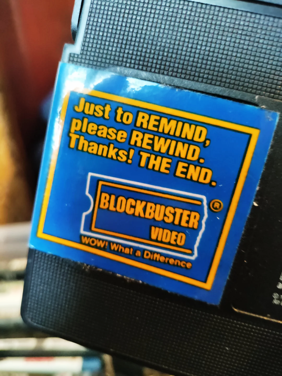 A close-up of a Blockbuster Video sticker on a VHS tape, reading: "Just to REMIND, please REWIND. Thanks! THE END." The sticker is blue with yellow and white text and a Blockbuster ticket logo.