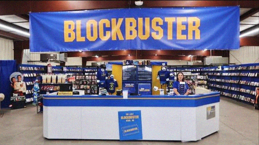 A Blockbuster video rental store with blue and yellow signage, shelves filled with DVDs and VHS tapes, and a counter where an employee stands, evoking a nostalgic atmosphere.