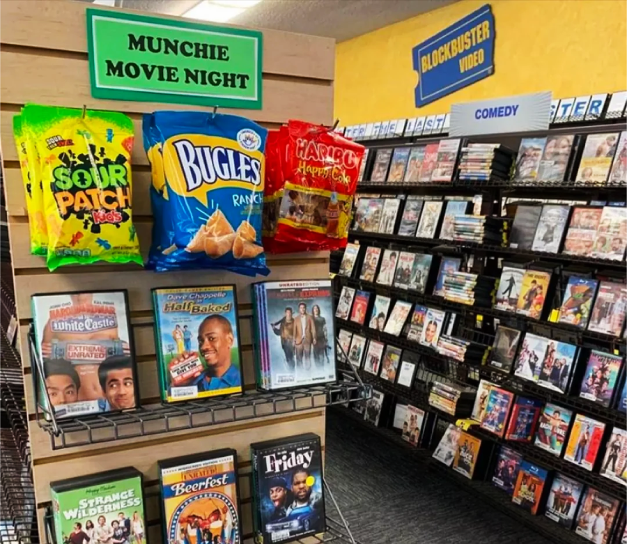 A Blockbuster Video store with shelves of DVDs, a "MUNCHIE MOVIE NIGHT" snack display featuring Sour Patch Kids, Bugles, and Haribo, plus classic movie cases like "Half Baked" and "Friday" below the snacks.