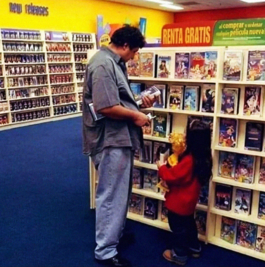 A man and a young girl stand by a shelf of DVDs or VHS tapes in a video rental store. The girl holds a stuffed toy. Shelves filled with more movies line the background, and signs advertise new releases and free rentals.