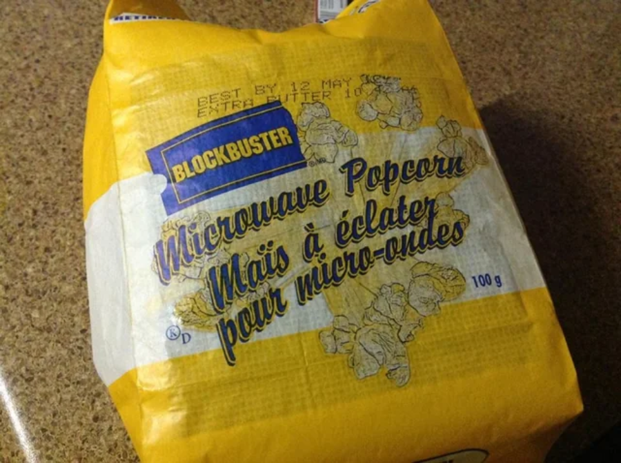 A yellow packet of Blockbuster microwave popcorn labeled in English and French, sitting on a brown countertop. The packaging features images of popped popcorn and an expiration date.