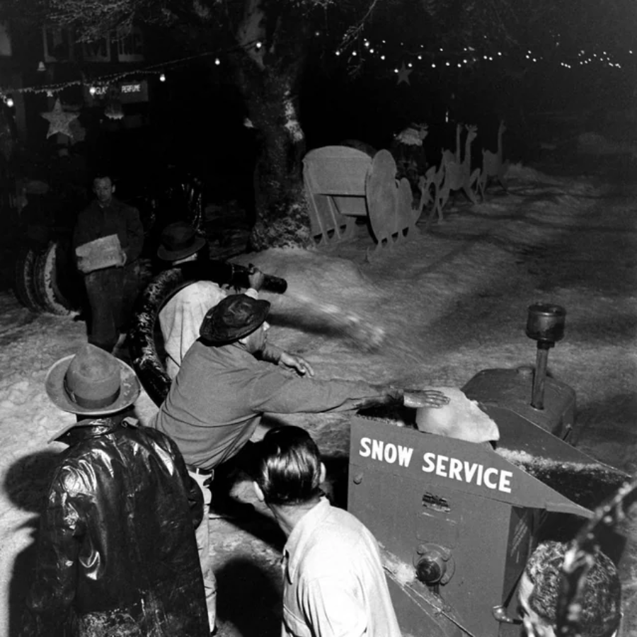 A group of people watch as a man uses a machine labeled "Snow Service" to blow artificial snow onto a snowy street decorated with Christmas ornaments at night.