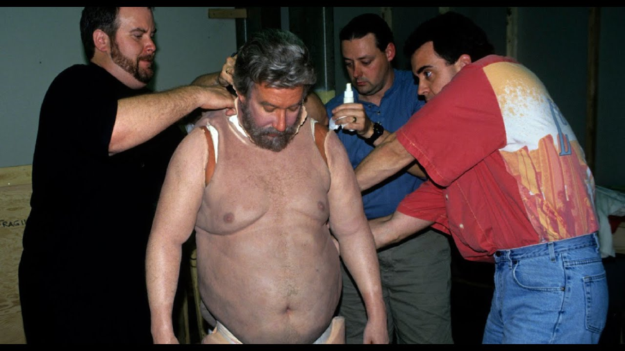 Four men are applying prosthetic makeup to a shirtless man with gray hair and a beard. The man stands still as the others work, attaching pieces to his shoulders and arms in a studio setting.
