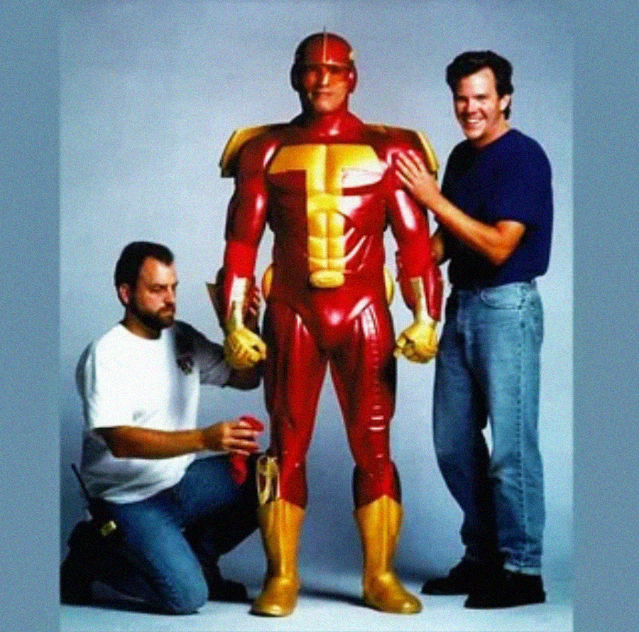 Two men pose with a person wearing a shiny red and yellow armored superhero suit. One man kneels while adjusting the costume's leg, and the other stands, smiling, with a hand on the suited person’s shoulder.