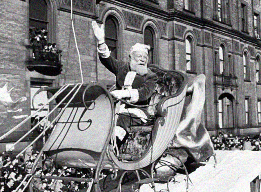 A man dressed as Santa Claus waves while sitting in a decorative sleigh during a festive parade, with a crowd and historic-looking building in the background.