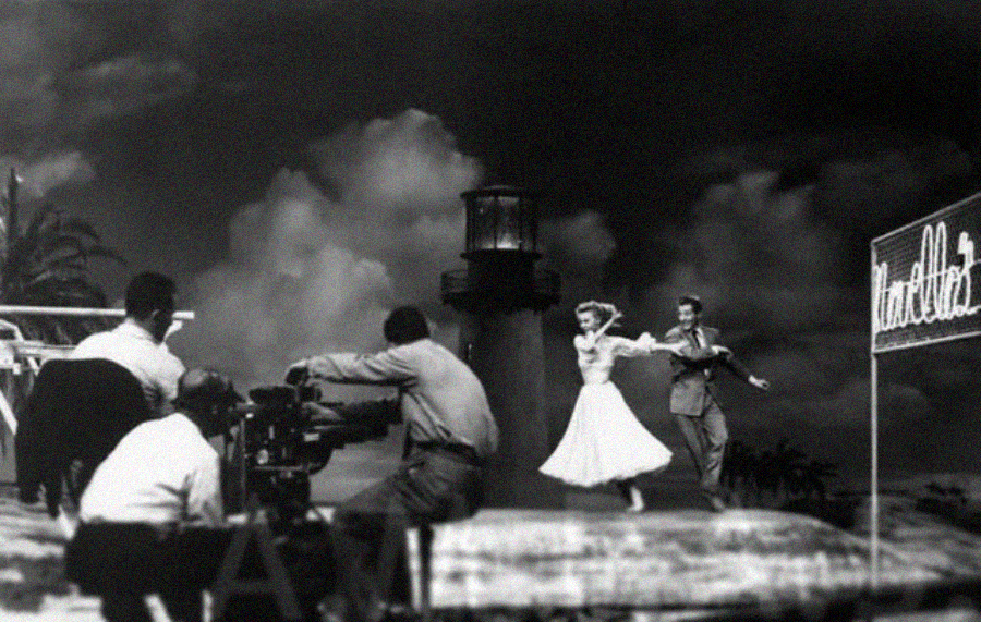 A black-and-white scene of a film set shows a camera crew filming a man and woman dancing in front of a lighthouse backdrop, with a large illuminated hotel sign to the right.