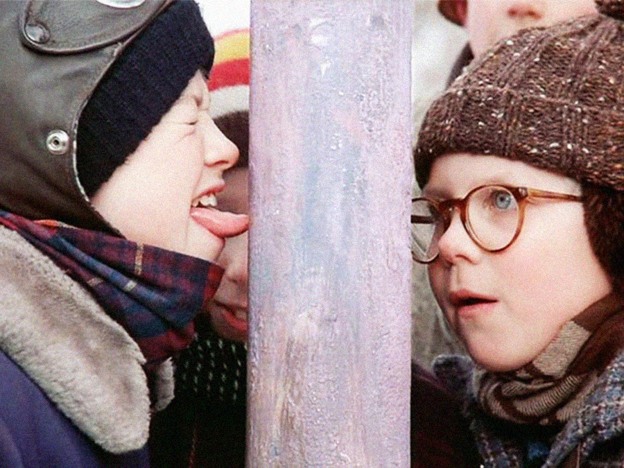 A child in winter clothes has his tongue stuck to a frozen metal pole while another child in glasses looks on with surprise; other bundled-up kids are partially visible in the background.
