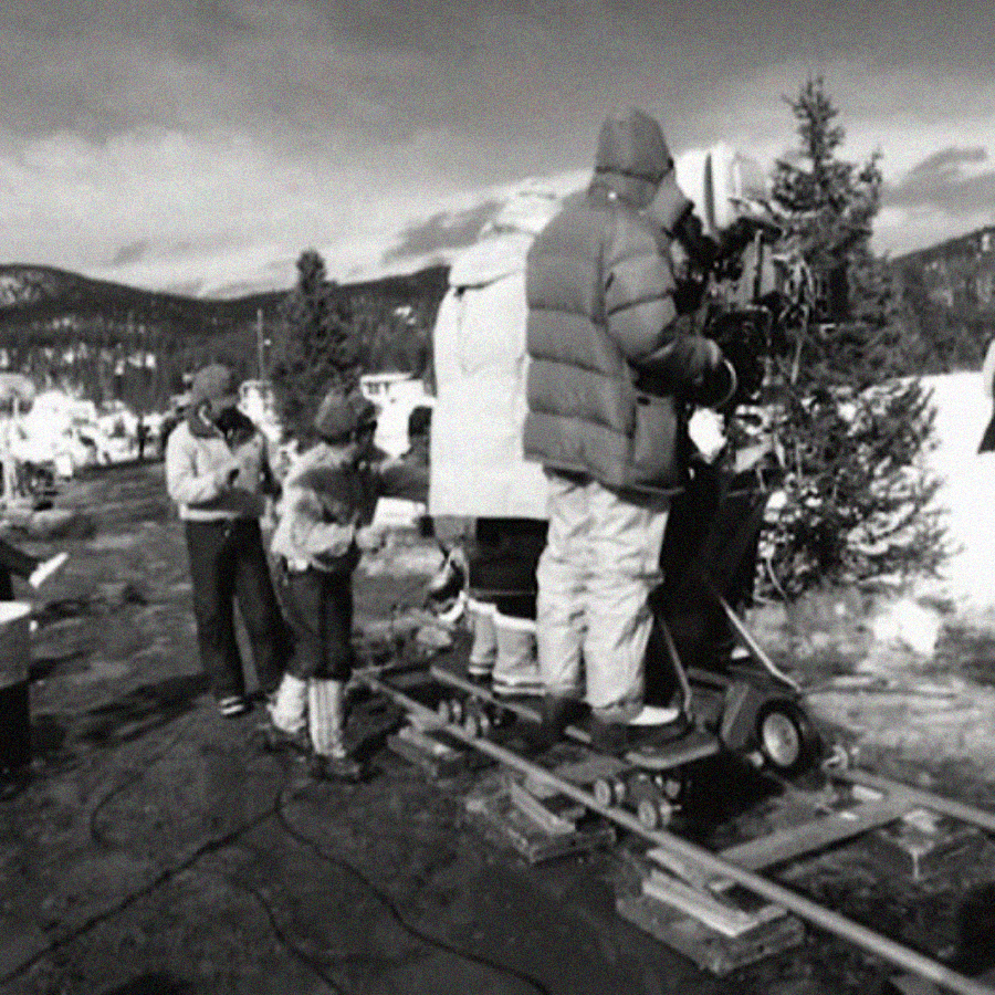 A black and white photo shows a film crew outdoors in a snowy, mountainous area. People wearing winter clothing operate a large camera on a dolly track, while others stand nearby among trees and snowbanks.