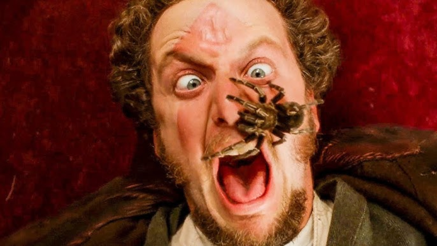 A man with curly hair screams in fear as a large spider crawls on his face, near his open mouth. His eyes are wide and he appears shocked, with a red background behind him.