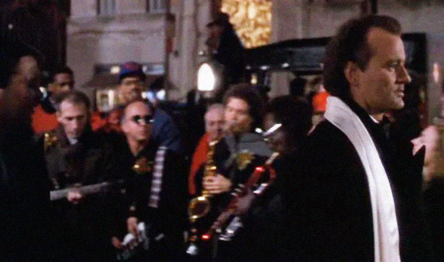 A man in a suit and white scarf walks past a group of musicians playing instruments, including a saxophone and guitar, in a lively, festive nighttime street scene.