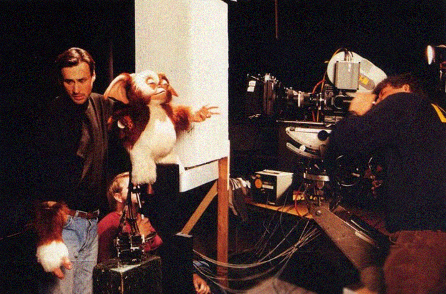 A puppeteer operates a large Gizmo puppet on a film set while another person manages a movie camera nearby, surrounded by film equipment and lighting.