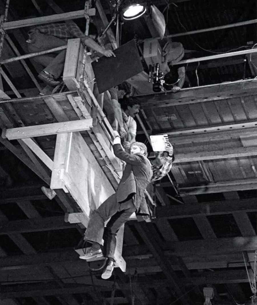 A man in a suit hangs from the edge of a wooden platform high above the ground while film crew members operate a camera and lighting equipment above him on a movie set.