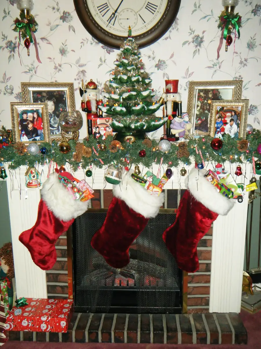 A fireplace decorated for Christmas with three red stockings, a garland with ornaments, a small Christmas tree, wrapped presents, framed photos, and nutcracker figurines on the mantel beneath a large clock.