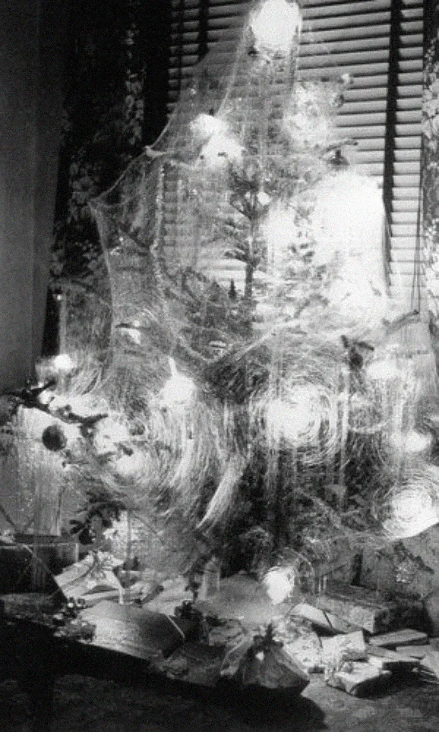 A vintage black-and-white photo of a Christmas tree decorated with tinsel and glowing lights, surrounded by wrapped presents, set in front of window blinds.