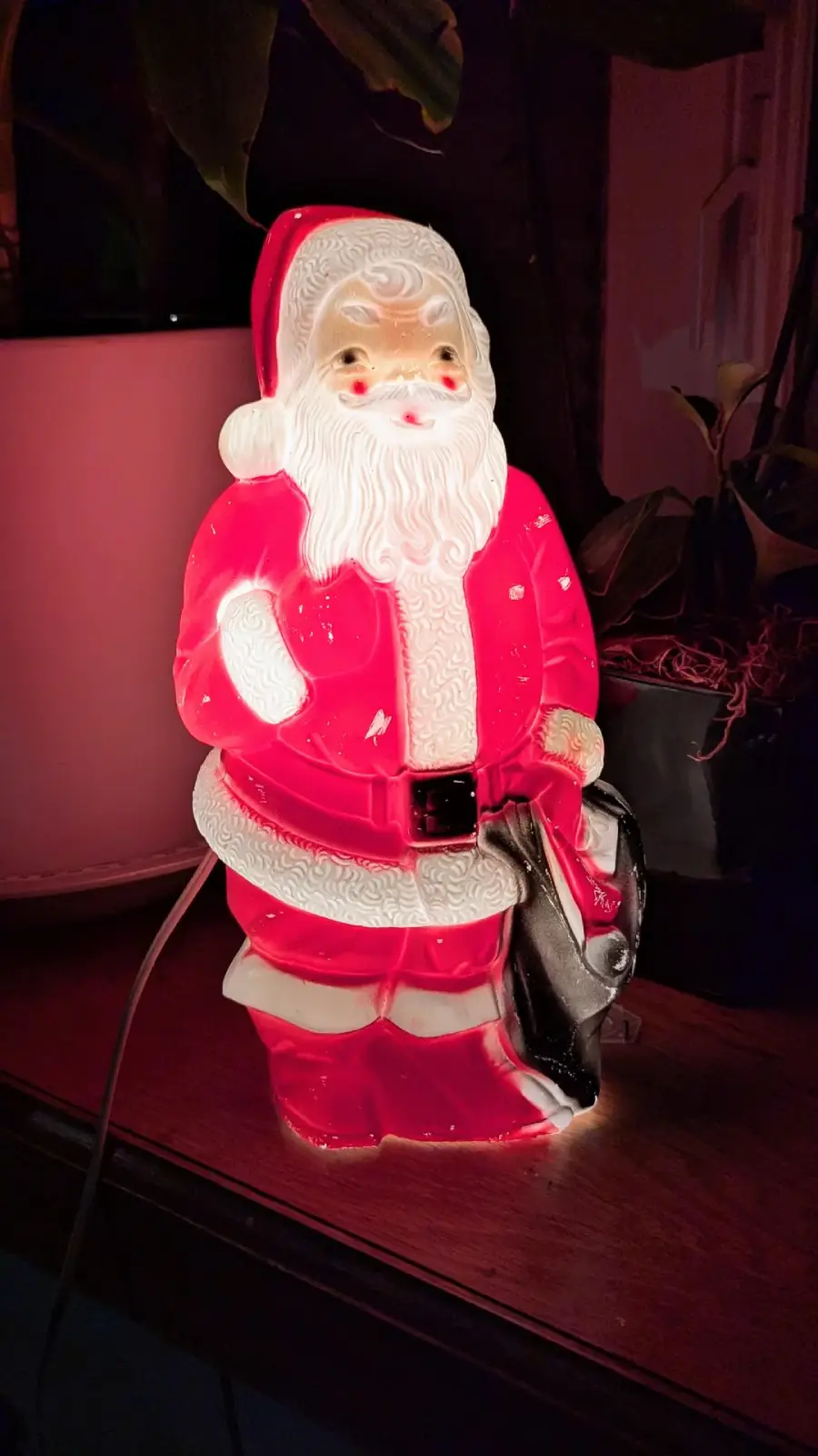 A glowing plastic Santa Claus figurine stands on a wooden surface, holding a black bag. The Santa is dressed in a traditional red suit with a white beard and hat, illuminated from within.