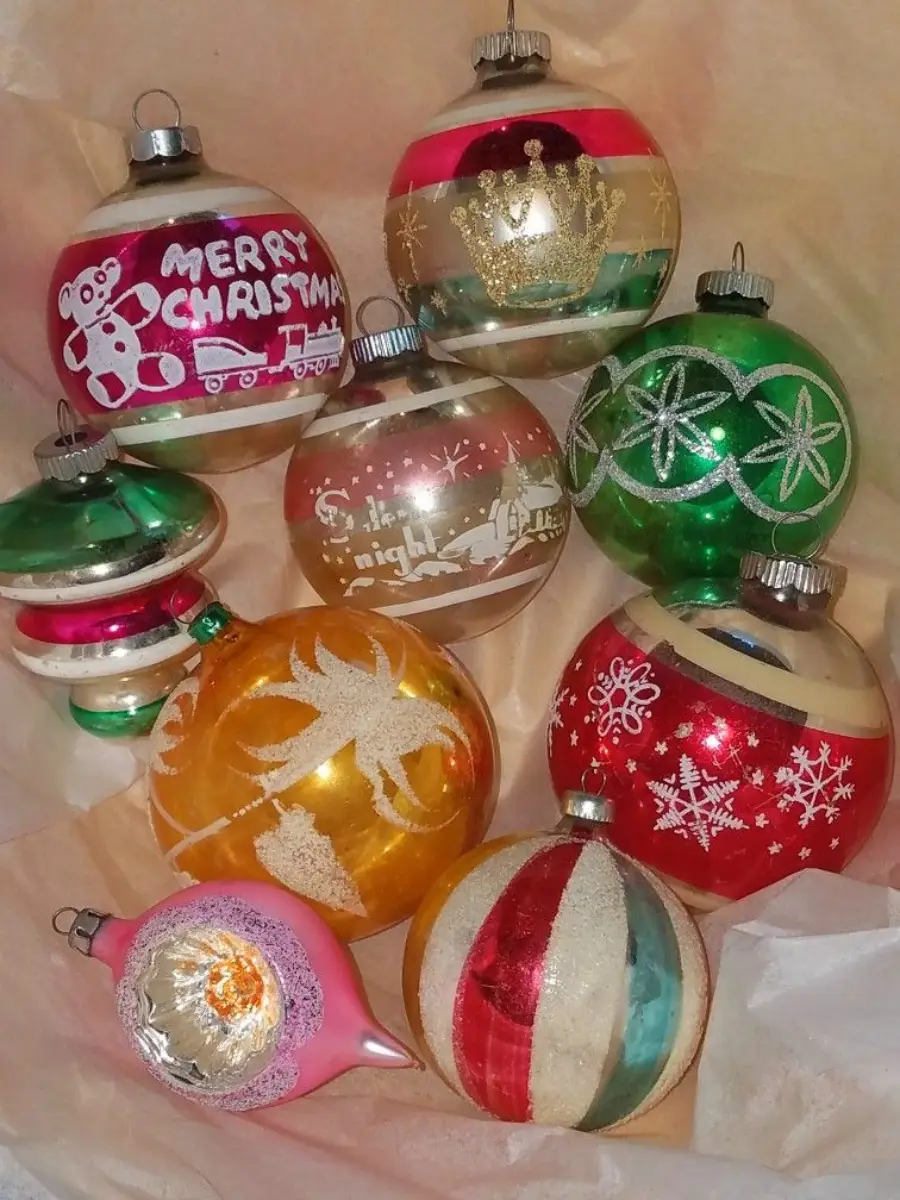 A collection of vintage glass Christmas ornaments with various shapes, colors, and festive designs, including snowflakes, a deer, stars, a crown, and one that says "Merry Christmas" with a teddy bear and train.