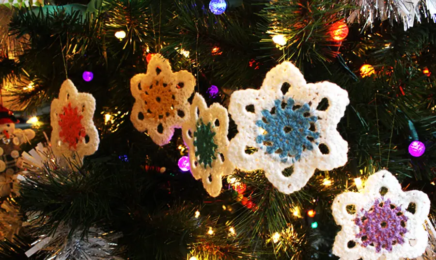 Crocheted snowflake ornaments in various colors hang on a Christmas tree decorated with lights, tinsel, and colorful baubles.
