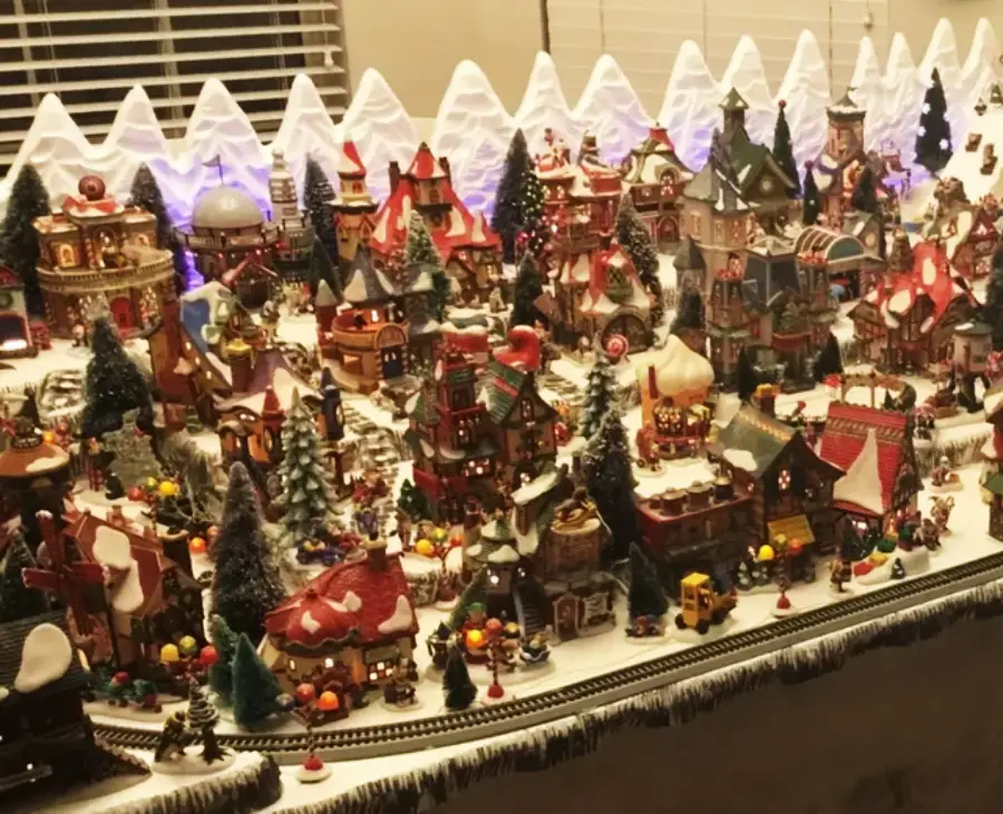 A detailed miniature Christmas village display with snow-covered buildings, trees, festive lights, and tiny figurines, set on a snowy landscape with a toy train running around the edge.