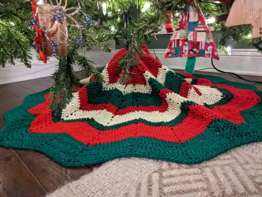 A crocheted Christmas tree skirt in red, green, and white covers the floor under a decorated Christmas tree with ornaments hanging above.
