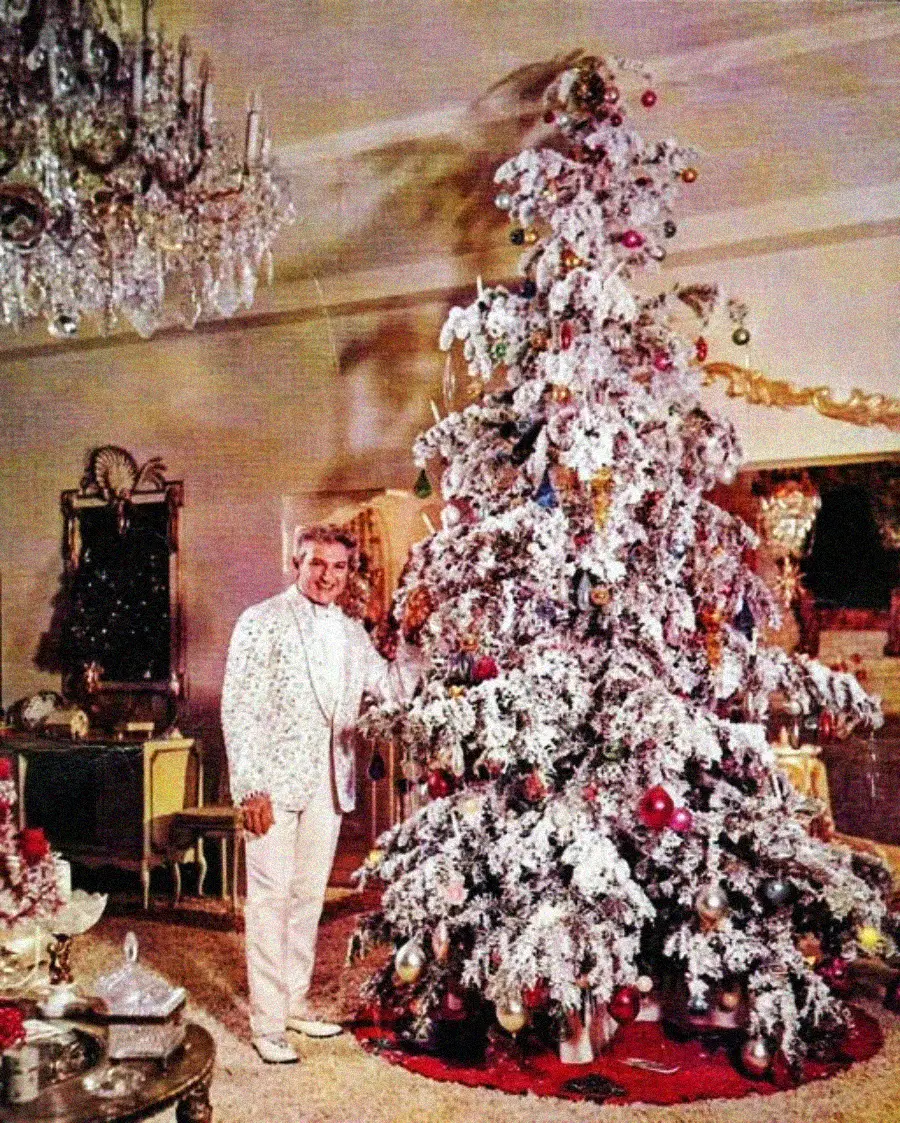 A smiling man in a white suit stands beside a large, heavily flocked Christmas tree decorated with various ornaments in an ornate, warmly lit living room.