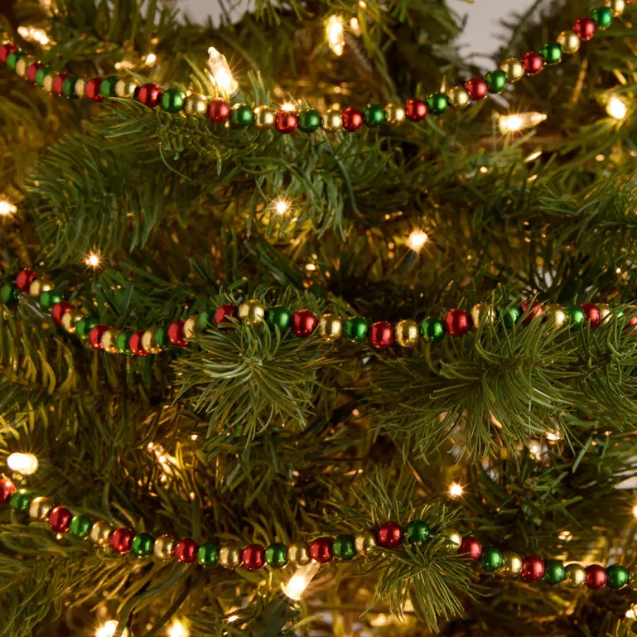 A close-up of a Christmas tree decorated with small string lights and a garland made of red, green, and gold beads. The tree branches are lush and green, creating a festive holiday atmosphere.