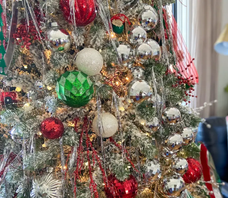 A close-up of a Christmas tree decorated with red, green, white, and silver ornaments, tinsel, string lights, and artificial snow on the branches.