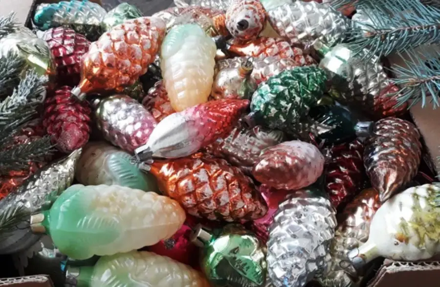 A box filled with colorful, vintage glass Christmas ornaments shaped like pine cones, with various shades of green, silver, gold, red, and pink, some attached to pine branches.