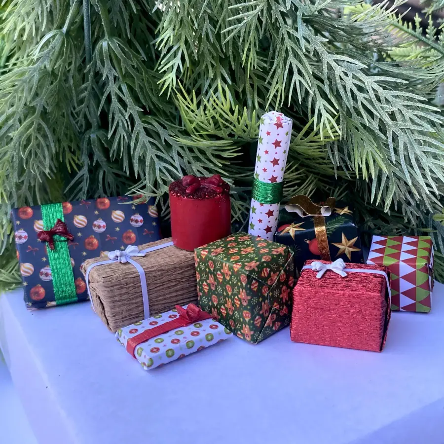 A variety of small, colorful gift boxes with festive wrapping and ribbons are arranged in front of green, pine-like foliage on a white surface.
