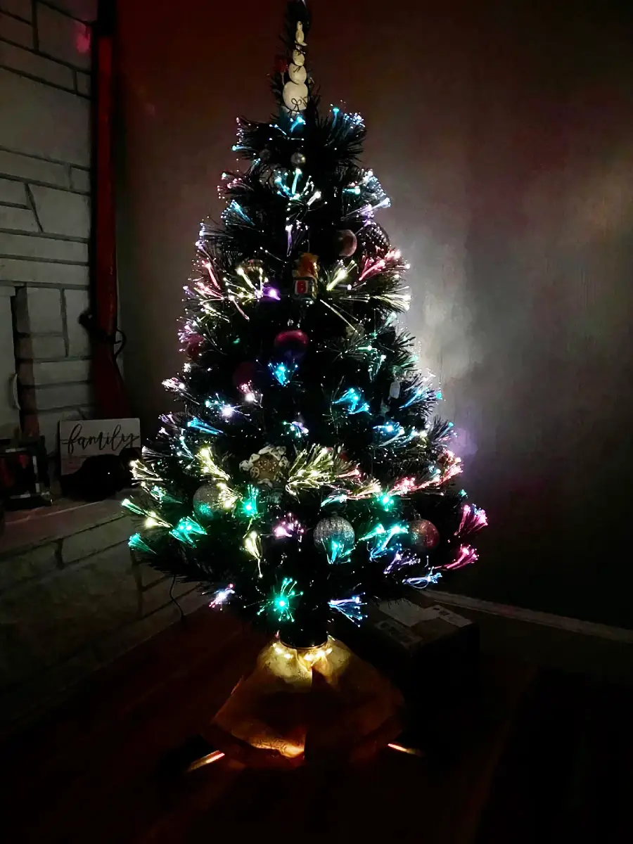 A Christmas tree decorated with colorful, glowing lights and ornaments stands indoors near a fireplace, with a golden tree skirt at the base and presents partially visible underneath.