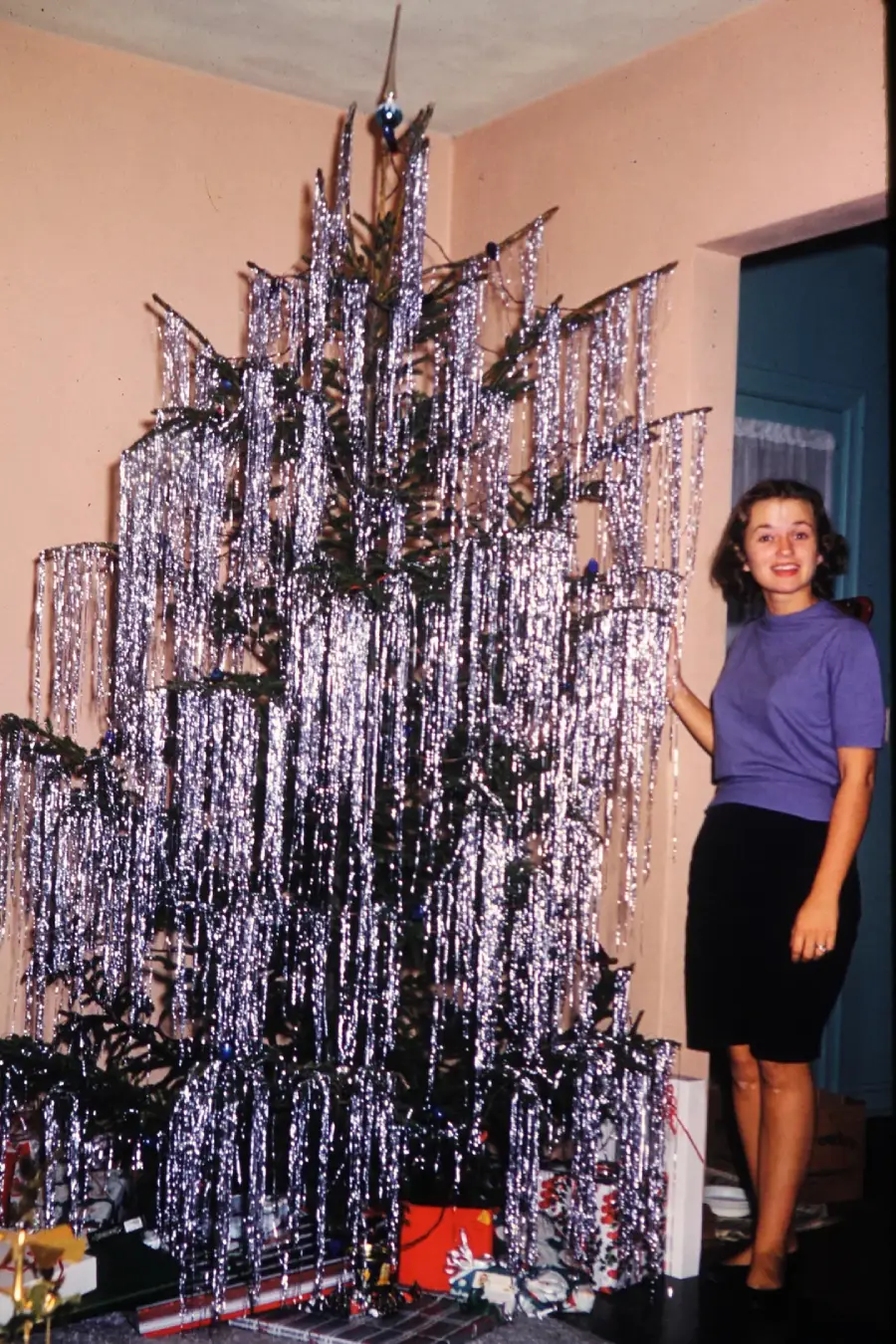 A woman stands beside a Christmas tree covered in shiny silver tinsel, with a few presents underneath. She is wearing a purple top and black skirt, and smiles at the camera in a warmly lit room.