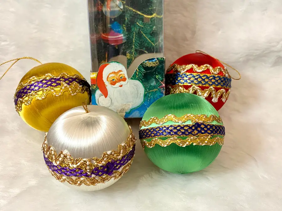 Four colorful Christmas ornaments decorated with gold and purple ribbon are arranged on a white surface. Behind them is a box featuring a festive image of Santa Claus and a decorated tree.