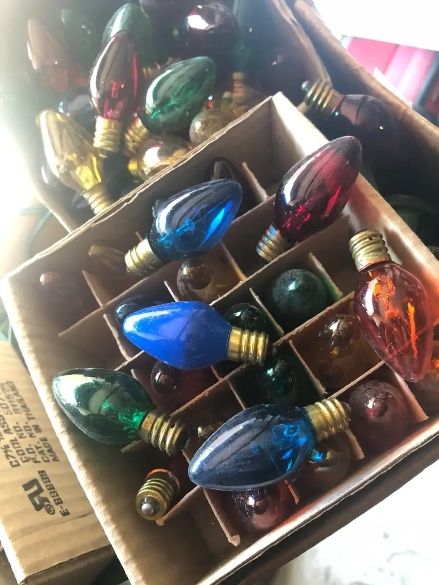 A close-up of a box filled with vintage, multicolored Christmas light bulbs, each in its own cardboard compartment. Bulbs are red, green, blue, and amber, with a few visible light scratches and wear.