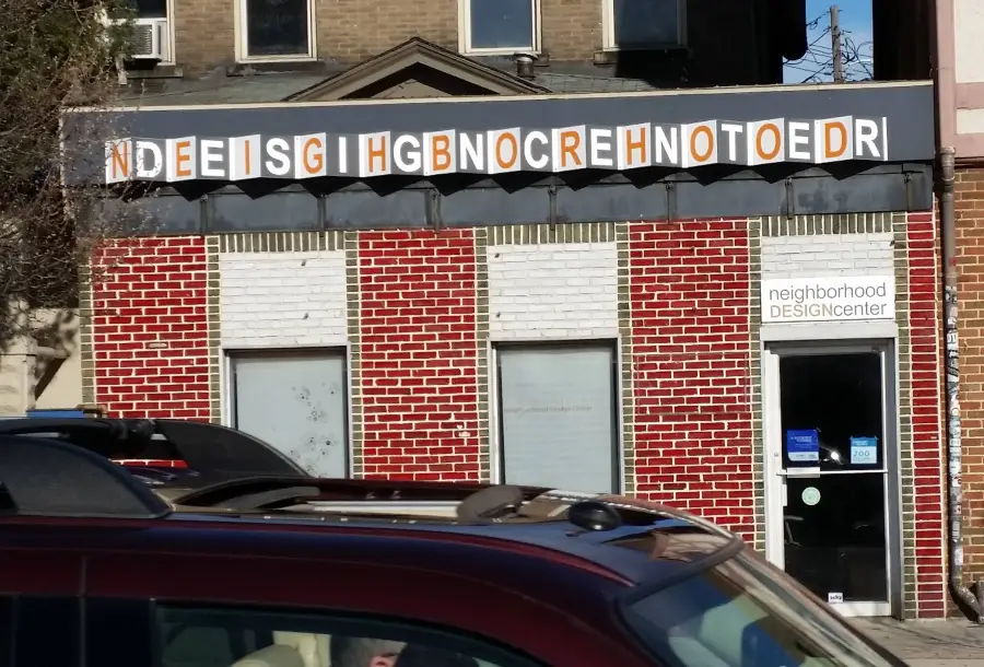 A brick building has a sign with jumbled letters above the windows reading "NDEEISG IHGBNOCREHNOTOEDR." A door on the right has a sign for "neighborhood DESIGNcenter." Cars are parked in front.