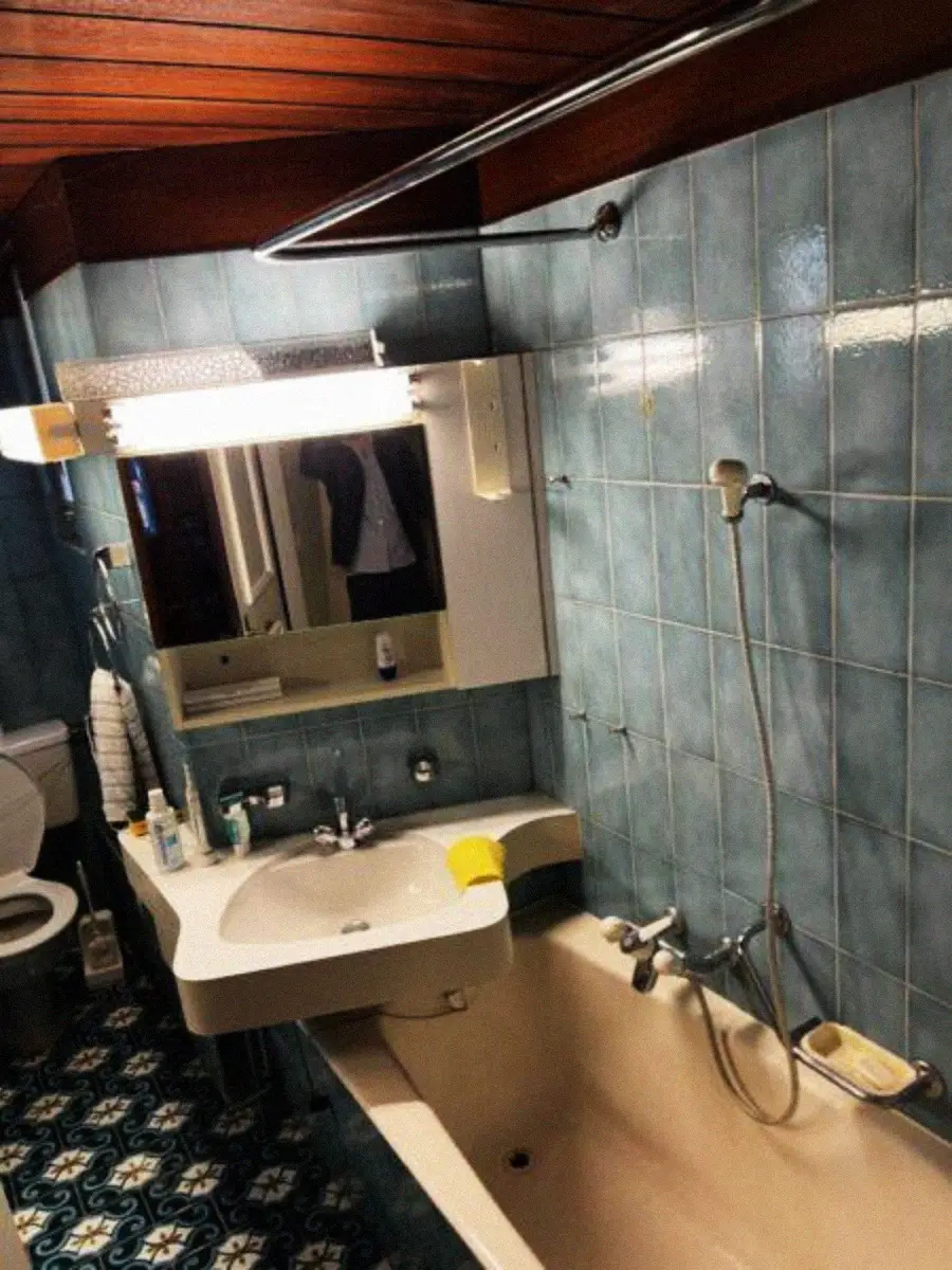 A vintage bathroom with blue tiled walls, a bathtub with a handheld shower, a white sink, a mirror above the sink, a toilet, and patterned floor tiles. Various toiletries are on the sink and bathtub edge.