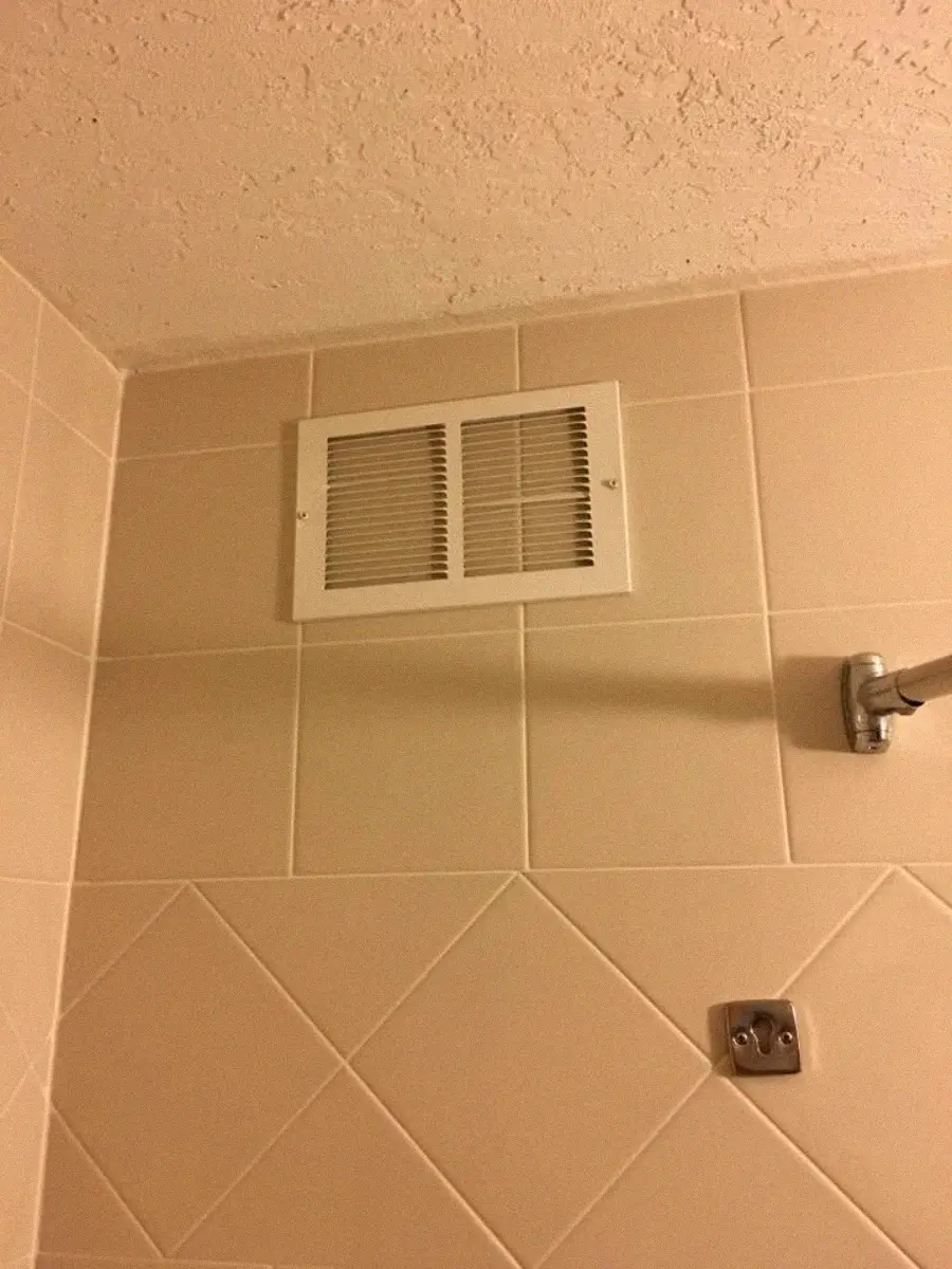 A beige bathroom wall with square tiles, featuring a white air vent near the ceiling, a metal shower rod extending from the right, and a metal fixture mounted on the wall below the vent.