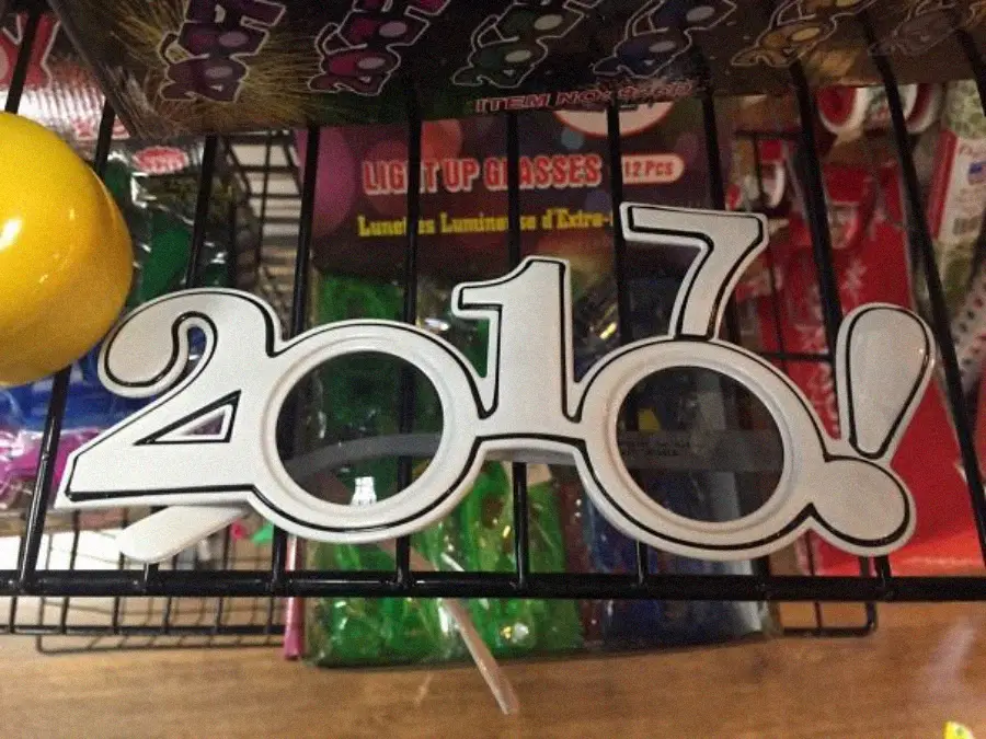 Novelty glasses shaped like the number 2017 are displayed on a store shelf. The glasses have large, round openings for eyes inside the zeros. Brightly colored party supplies and packaging are visible in the background.