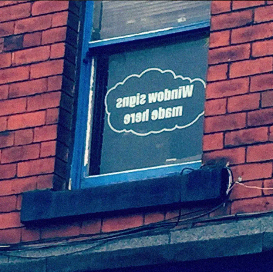 A sign in a window reads "Window Signs made here," but the text is reversed, as seen from outside a brick building.